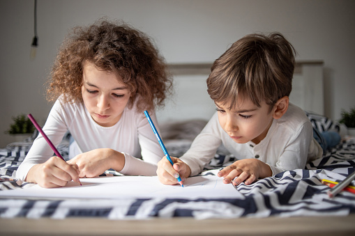 Two children, brother and sister, draw on paper with wooden crayons while on the bed in the bedroom, carefree childhood and free time