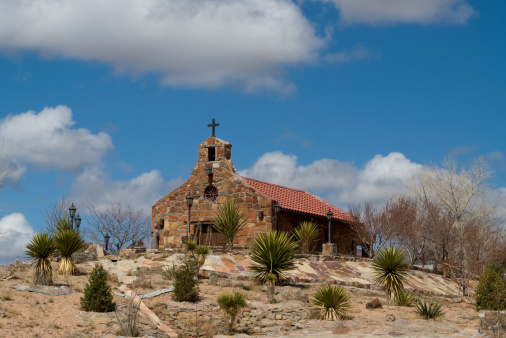 This is a calvary style chapel, with rock walls and a spanish tile roof.  The chapel sits on a small knoll and is surrounded by yucca trees.  Overhead is a sky of deset blue, with puffy white clouds.