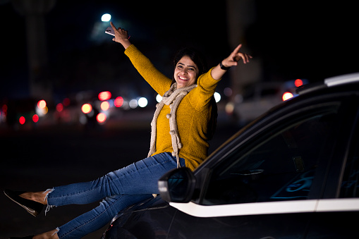 Carefree young woman sitting on car hood with her arms raised in excitement outdoors at night