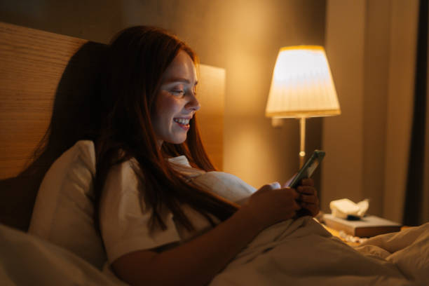 istock Side view of cheerful redhead young woman using smartphone, looking on screen, typing online message on social media, lying on bed late at night. 1470261124