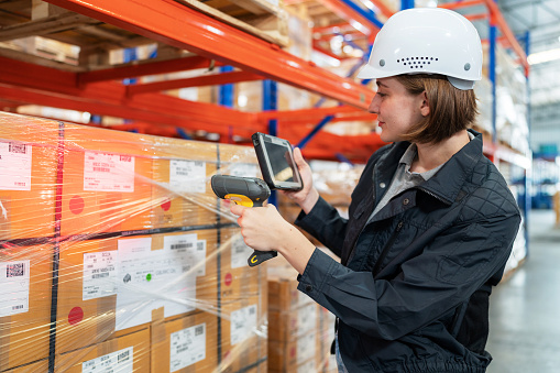 Implementing RFID tags help make delivery easier and growing up your business sustainably. Caucasian warehouse worker with safety helmet using a barcode reader to scan QR code on the box standing in the distribution warehouse. Looking at tablet for checking stock inventory.