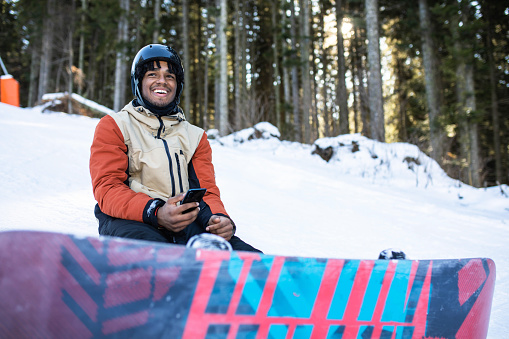 A multiracial snowboarder is using his phone on the slopes while taking a break from snowboarding