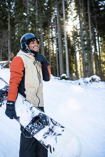 A handsome multiracial man is smiling while carrying a snowboard at a ski resort
