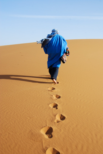 A Berber man walks barefoot in the sand of the Sahara Desert in Morocco leaving footprints behind.
