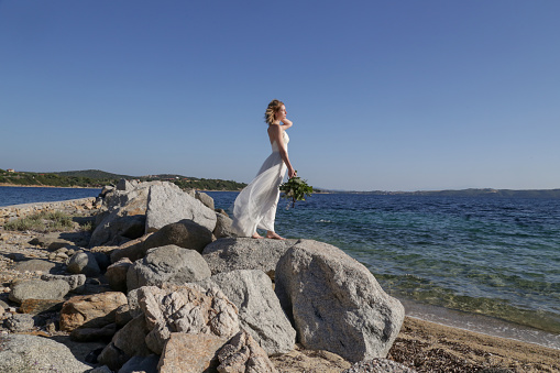 Bride in long white wedding dress standing on windy sea rock shore holding lush green wedding  bouquet with boho twist