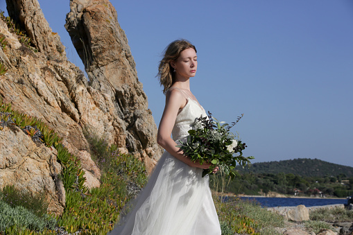 Bride in long white wedding dress standing on windy sea rock shore holding lush green wedding  bouquet with boho twist