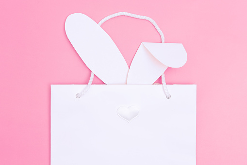 Shopping for Easter. White paper shopping bag with white bunny ears on pink background. Concept Easter gifts, preparation, holiday surprise.