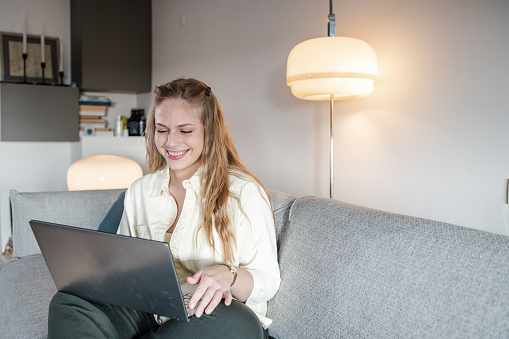 Young woman, with a long blond hair, relaxing at home, smiling and looking at a laptop.