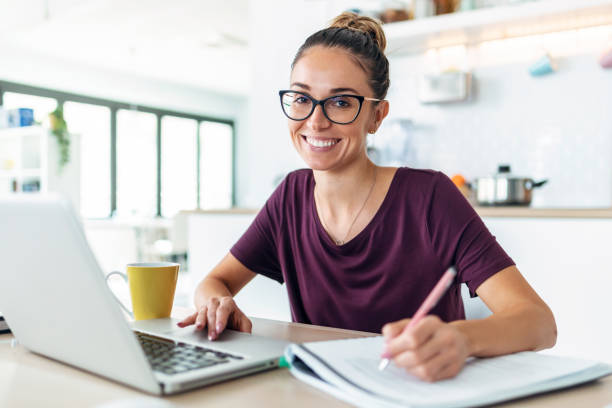 istock Pretty young woman working with laptop and documents while looking at camera in the kitchen at home. 1470308494