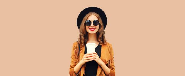 Portrait of beautiful young woman with cup of coffee wearing black round hat on brown background stock photo