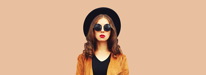Portrait of beautiful stylish young woman wearing black round hat, jacket on brown background