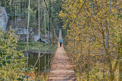 Senior woman with gray hair in casual clothes walking on suspension bridge on background of nature. Back view in full length. Outdoor activities concept