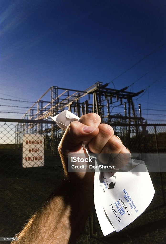 Electric Shock Clenched fist of man holding an electric bill for $14,300 in front of power substation Consumerism Stock Photo