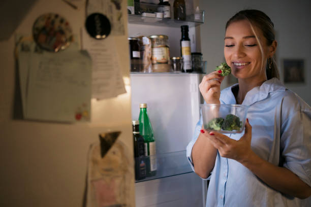 Young woman enjoying eating broccoli by the open fridge at night in kitchen stock photo