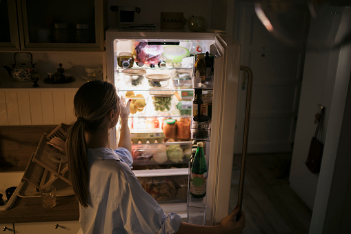 Rear view of a woman opening refrigerator in middle of night and looking what to eat at home. Young woman looking into fridge at night in kitchen.