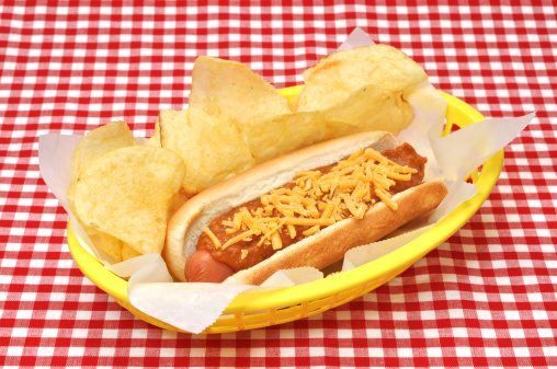 Hot dog with chili, cheese, and potato chips in basket on red gingham tablecloth.