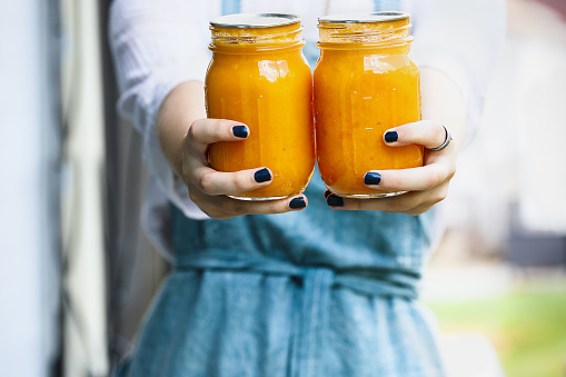 Woman in an apron holding out fresh homemade peach butter. Home preservation. Selective focus on mason jar with blurred background.