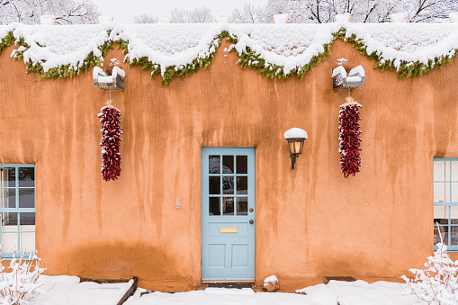 Christmas scene in Santa Fe, New Mexico, snow-covered adobe building with red chile ristras, green garland, and glowing light