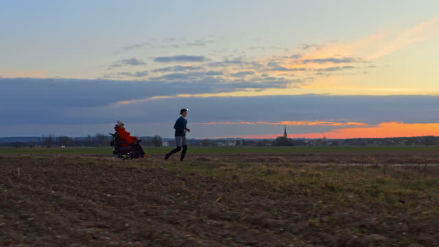 AERIAL Young Girl in Wheelchair Joins Brother on Scenic Sunset Jog through Country Fields