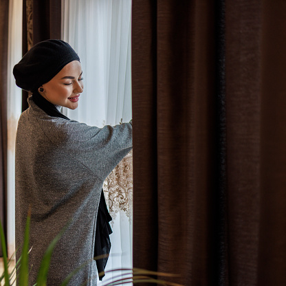 Young beautiful Muslim woman is opening blinds on a window in a living room.