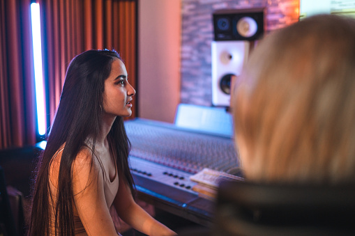 Young Hispanic smiling female singer talking with the senior Caucasian male composer in the recording studio. Waist-up, all looking away.