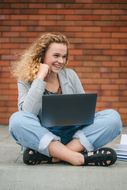 smiling curly-haired student girl sitting on street working on laptop, preparing for exams outdoors at university campus. technology and education concept. internet communication, wireless connection technology. - surfing wireless vertical outdoors lifestyles imagens e fotografias de stock