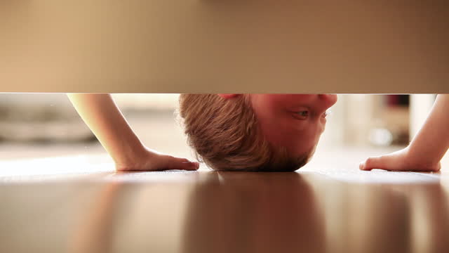 Cheerful little boy looking for something under the bed and sincerely smiling in the kids room. Happiness and carefully childhood concept video.