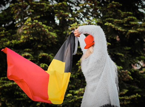 Close-up side view portrait of a beautiful blonde middle-aged Caucasian woman wearing a hat on her head waving the Belgian flag for Independence Day in the backyard of her house. Belgium day concept, flags, symbols, celebration, football, moms, fans.