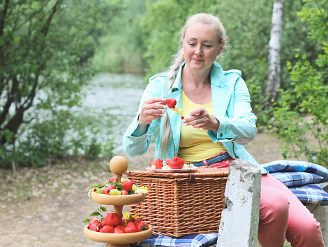 Beautiful middle aged caucasian blond girl happy with a smile preparing fruit skewers of strawberries, grapes and cheese, dressing them on wooden skewers, sitting on benches with a wicker basket in the forest, close-up side view side view. The concept of a picnic, outdoor recreation.
