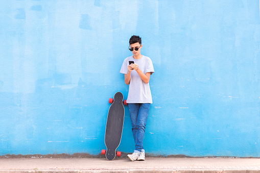 Man standing on the sidewalk of the street and leaning against a blue wall while using his smartphone to chat online on social media, with a longboard on the side.