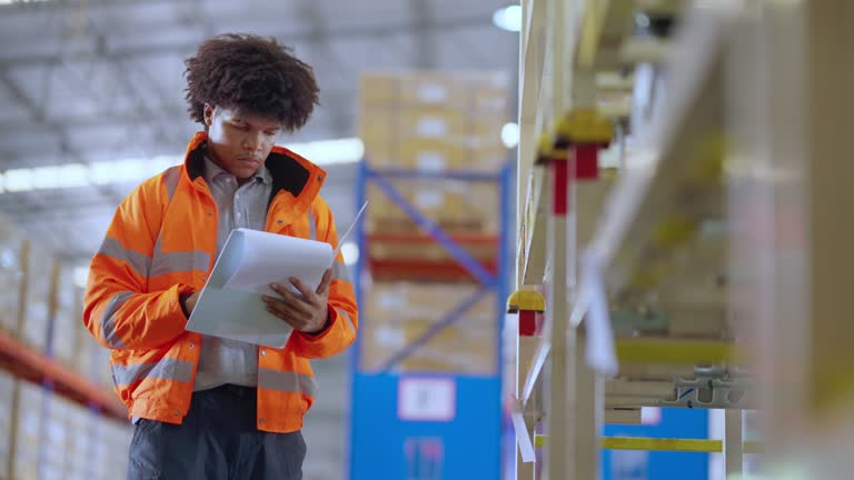 Data comparerison into Warehouse Operations. African warehouse worker checking on a clipboard and compare data of number of parts to review inventory balance data while standing by a shelf rack in a distribution warehouse.