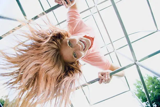 A low angle view of a young woman hanging from monkey bars on a playground and smiling at the camera. Horizontal shot.
