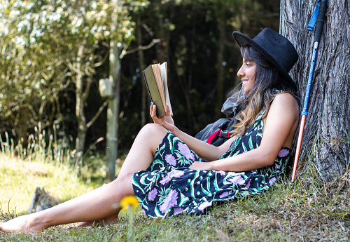 A woman in a black dress and hat sits under a tree reading a book on a sunny day in the countryside