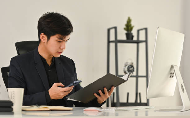 istock Smart young businessman holding open office binder with paperwork and using smart phone at workplace. 1470417778