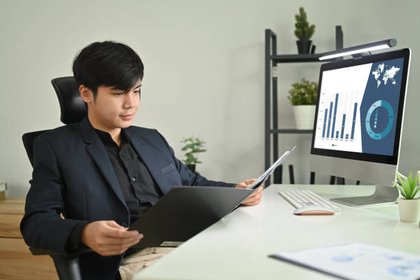 istock Confident asian male accountant advisor holding open office binder with paperwork while sitting at workstation. 1470417802