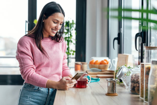 istock Beautiful woman using her mobile phone while drinking a cup of tea in the kitchen at home. 1470427442