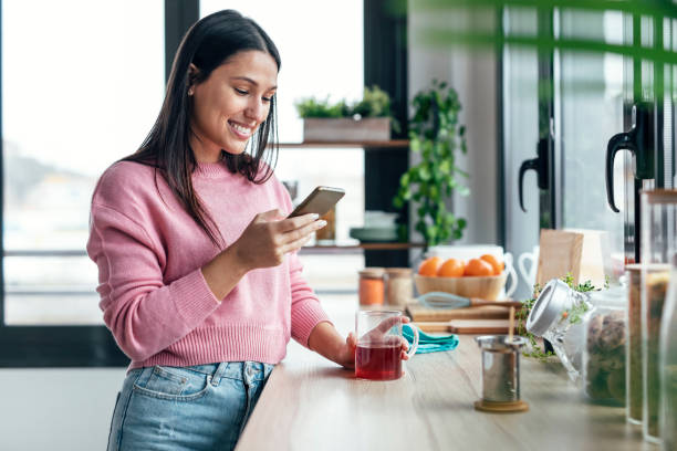 istock Beautiful woman using her mobile phone while drinking a cup of tea in the kitchen at home. 1470427457