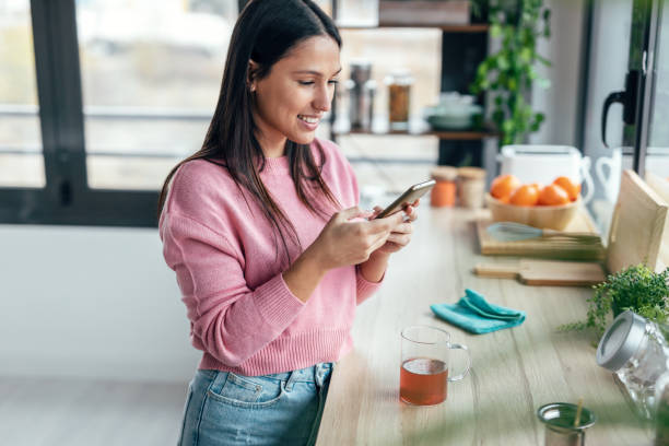 istock Beautiful woman using her mobile phone while drinking a cup of tea in the kitchen at home. 1470427461