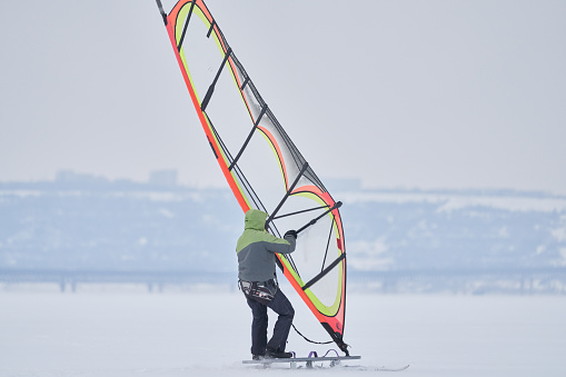 A middle-aged man, a snowsurfer, rides a sailboard on a snow-covered frozen lake. Snowsurfing on a cloudy winter day.