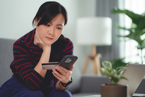 Asian woman holding credit card using smartphone have a problem with shopping and paying online on website sitting on sofa in living room at home.