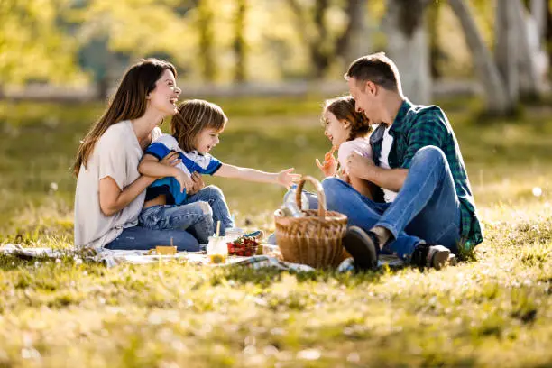 Cheerful parents and their small kids having fun while talking during springtime picnic in nature.