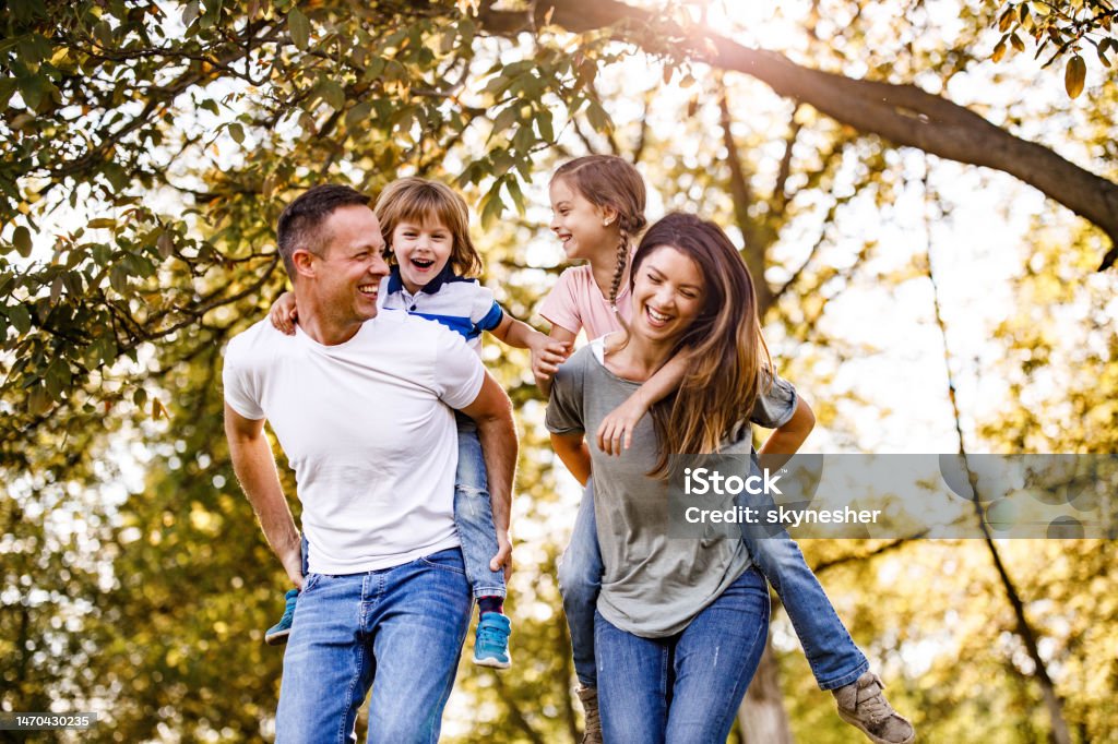 Carefree parents and their small kids having fun while piggybacking in springtime at the park. Young happy family having fun while piggybacking in spring day. Mother Stock Photo