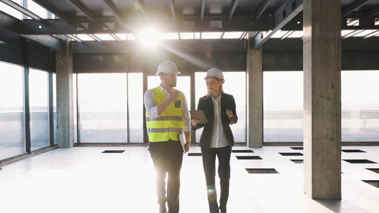 Workers Partners Holding Drawing Plan and walk at Modern Unfinished Business Center. Male Civil Engineer and Young Female Building Architect Discussing Project Work on a City Construction Site.