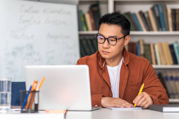 Korean Middle Aged Teacher Using Laptop Taking Notes In Classroom stock photo