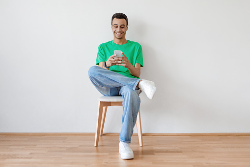 Excited young arab man using cellphone and texting or networking in social media sitting on chair over light wall. Modern communication, gadgets and technology