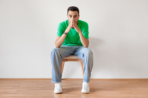 Negative emotions. Young arab guy with thoughtful face expression, feeling sad, having problem, suffering from depression or stress, sitting on chair against light studio wall