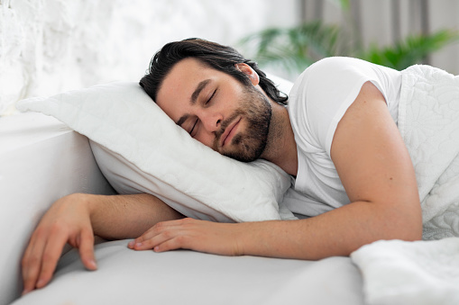 Healthy sleep concept. Closeup of handsome bearded young man sleeping in nice comfortable bed at home, enjoying good sleep, side view, copy space, white bedroom interior