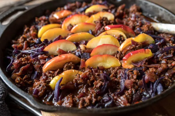 Ground beef stew with red cabbage or puple cabbage. Topped with butter fried apples and served hot and ready to eat in a cast iron pan on wooden table background. Closeup and front view. Traditional and rustic cuisine