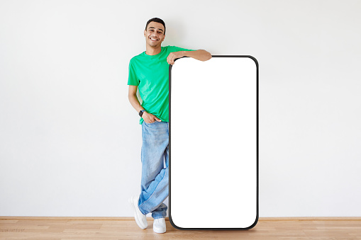 Online promo. Happy arab man standing near big blank smartphone demonstrating space for your ad, mockup. Guy leaning at mobile phone with white screen, full length shot