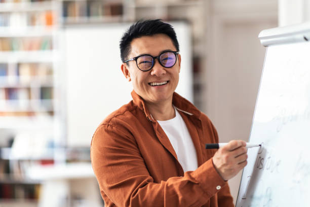 Positive Asian Teacher Man Smiling Writing On Whiteboard In Classroom Positive Asian Teacher Man Smiling To Camera Posing Writing English Grammar Rules On Whiteboard Having Distance Lesson In Classroom. Modern School Education And Knowledge Concept lecturer stock pictures, royalty-free photos & images